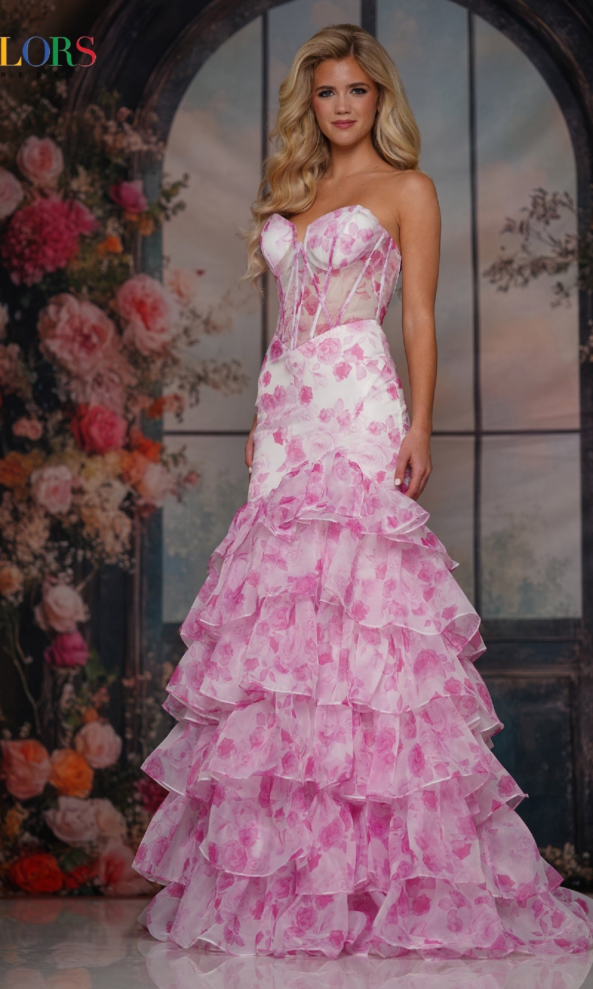 A woman wearing the Long Prom Dress: Colors Dress 3928, a strapless, ruffled pink and white floral gown, stands before a backdrop featuring large flowers and arched windows.